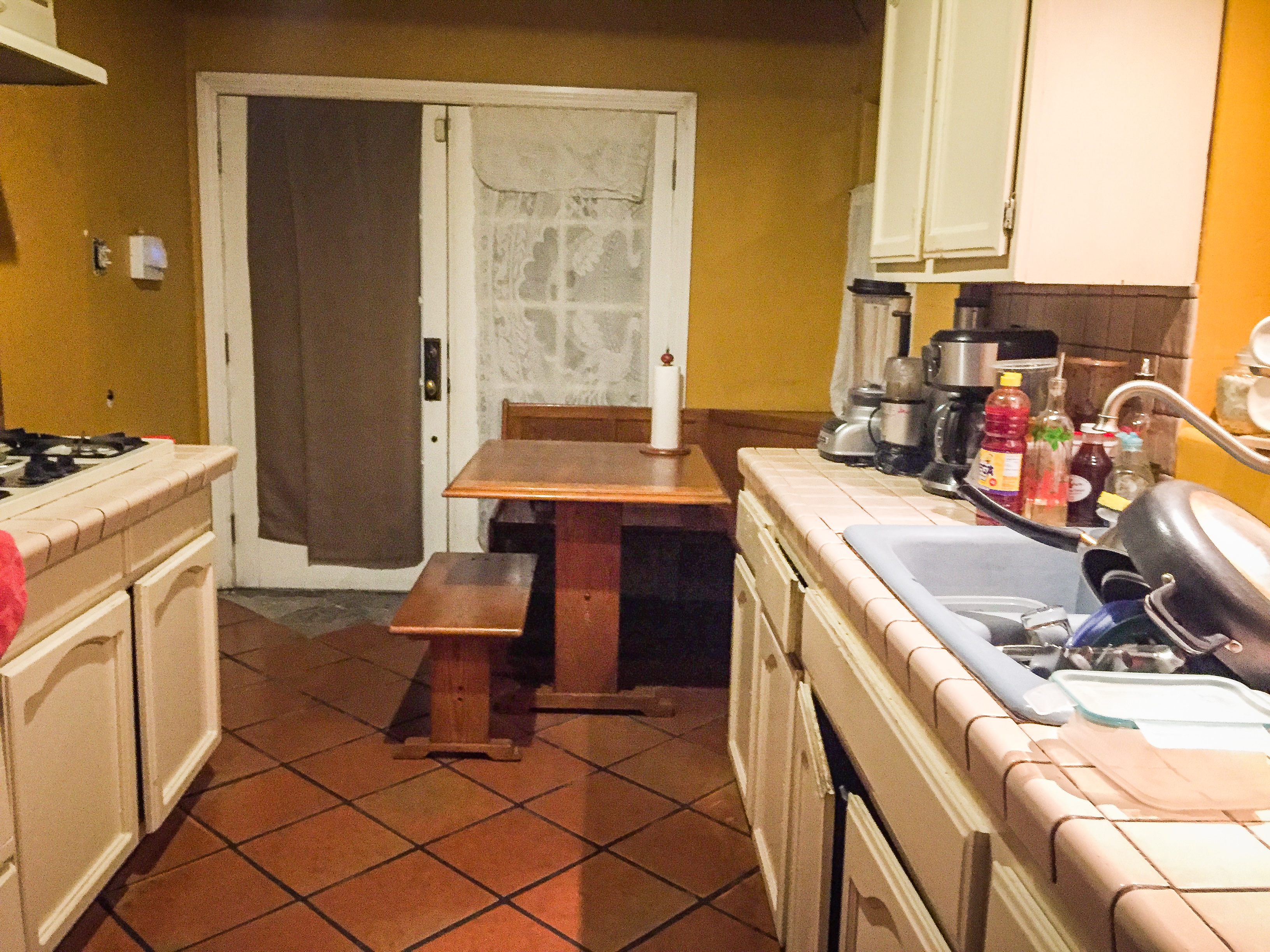 Original kitchen with dated tile counters, terracotta floor, and outdated cabinets